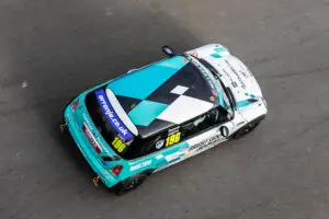 Aerial view of Ronnie Smith's MINI CHALLENGE race car, number 196, speeding on track. The car features a teal, white, and black livery with sponsor logos including Bright Signs, Astrasec, Millers Oils, and Oranje.co.uk. The name 'Ronnie Smith' is displayed on the side window.